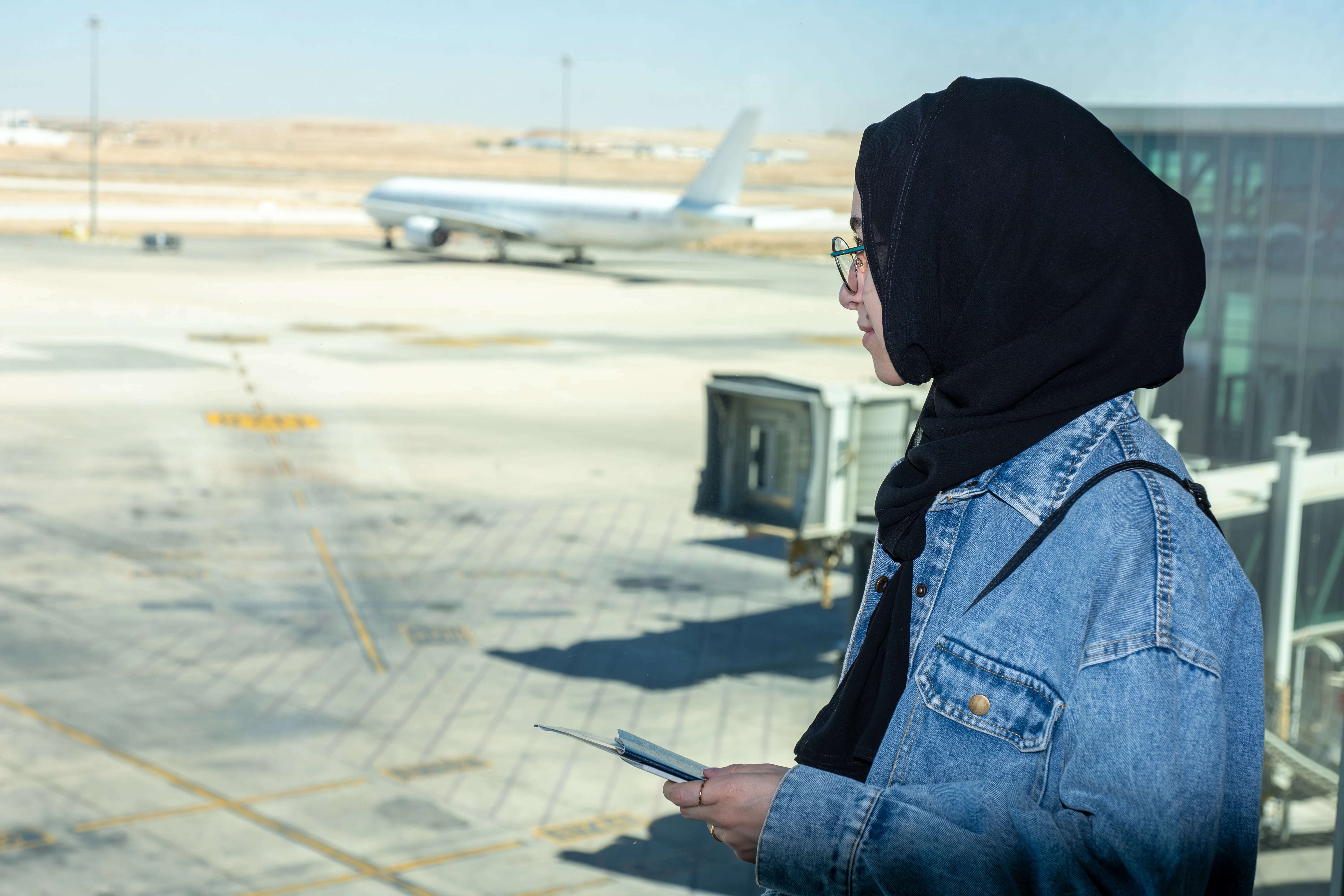 Young arab female holding her passport at the airport ready for new adventure with airplane in the background