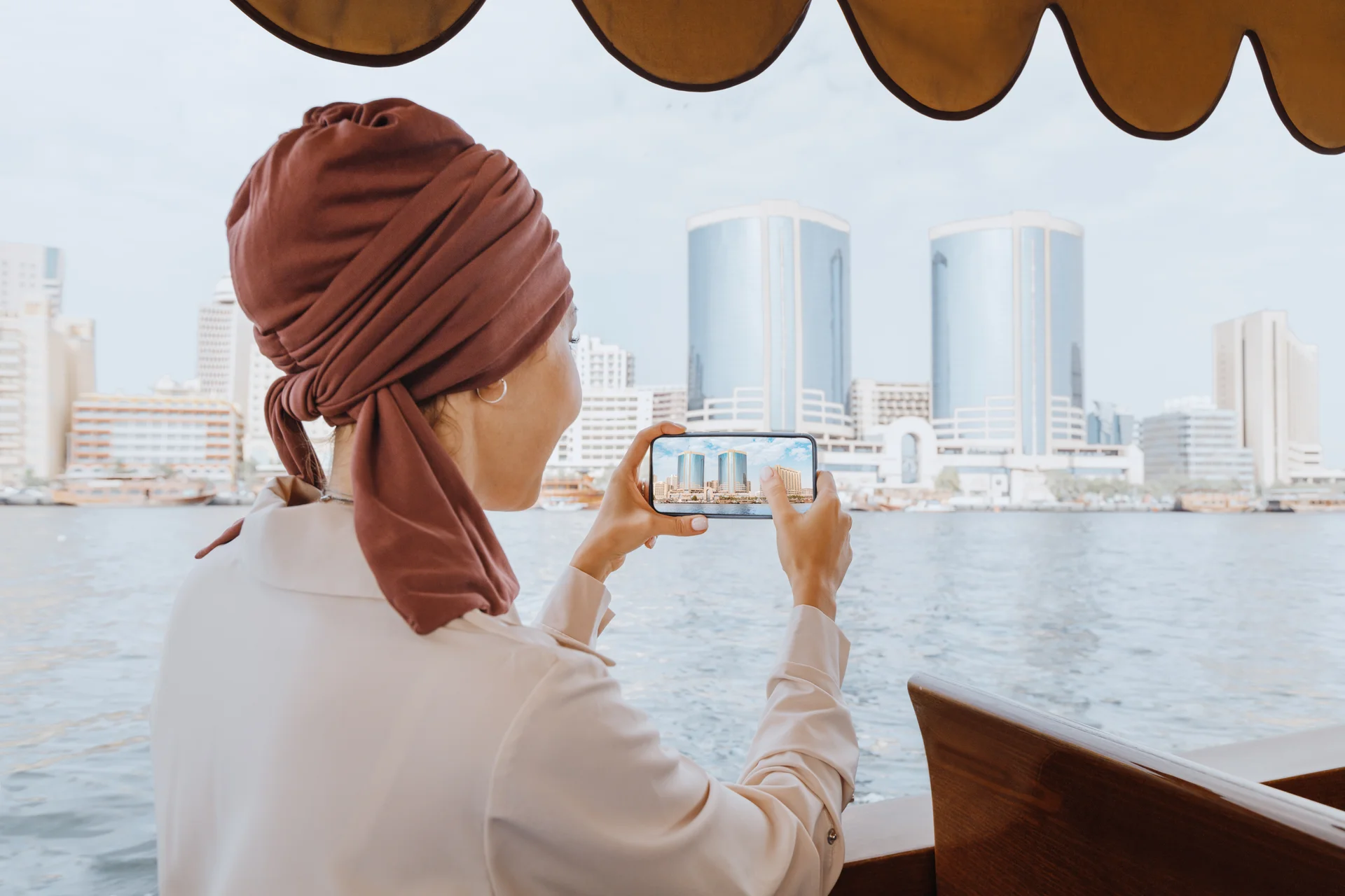 Happy asian woman in a red turban takes pictures on her smartphone while cruising on a traditional Abra Dhow boat on Dubai Creek Travel in UAE concept